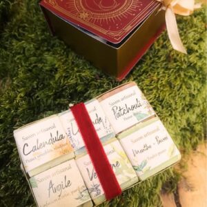 Un ensemble de petits savons artisanaux emballés dans du papier illustré est maintenu par un ruban rouge. Les savons portent des étiquettes indiquant différentes variétés comme Calendula, Argile, Patchouli, Verveine, Menthe-Poivrée. À côté, on voit une jolie boîte cadeau décorative rouge et dorée avec un ruban crème. L’ensemble est posé sur un tapis de mousse verte, créant une ambiance naturelle et chaleureuse.