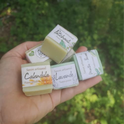 carré la masca Main tenant quatre savons artisanaux La Masca : Calendula, Lavande, Patchouli, et Menthe-Pinède. Savons naturels aux étiquettes colorées, photographiés en extérieur avec un fond végétal flou.