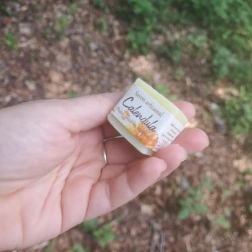 Main tenant un petit savon artisanal La Masca au calendula, adapté aux peaux sensibles. Étiquette illustrée d’une fleur de calendula, fond naturel flou avec feuilles et terre.
